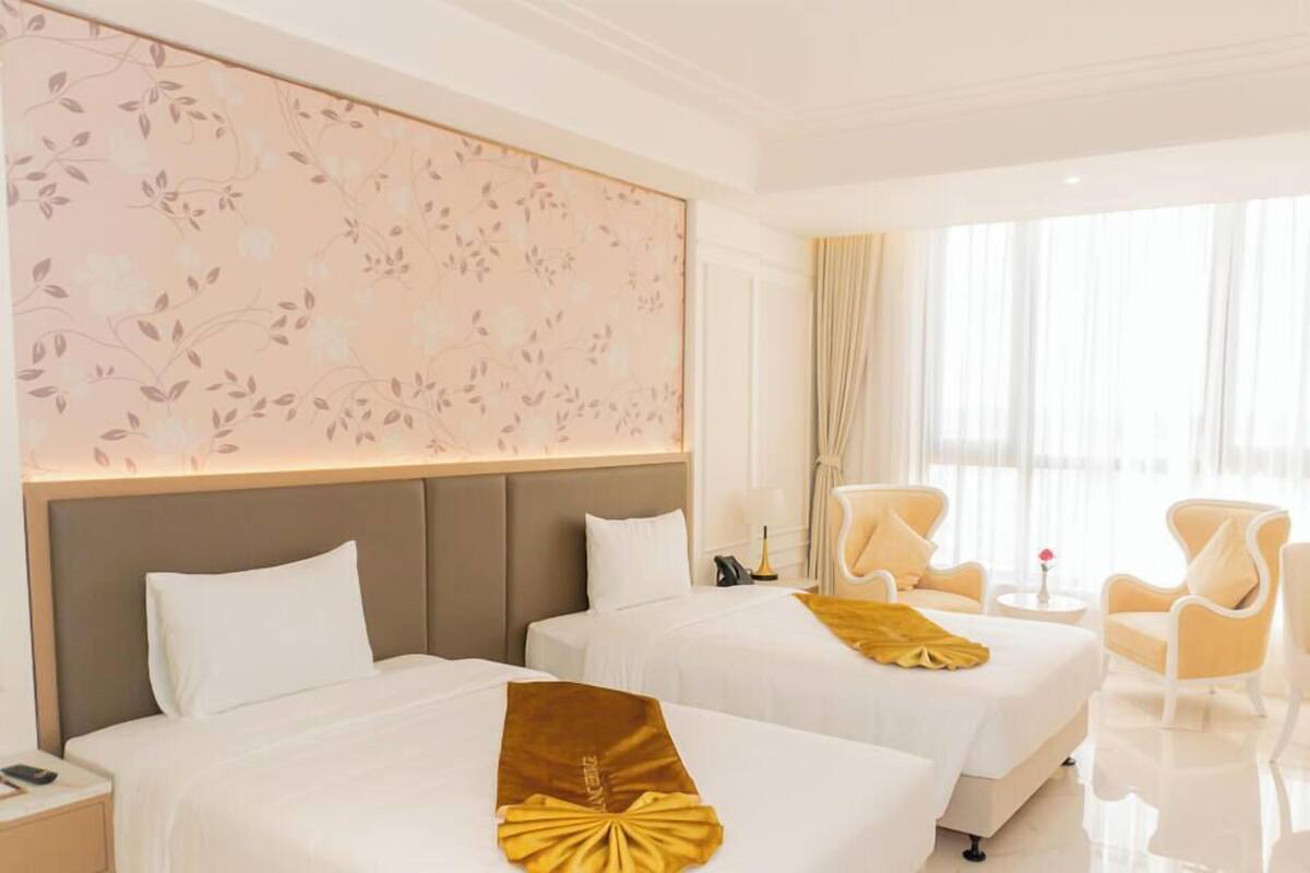 Habitación de hotel con dos camas, una pared con motivos florales, un rincón de estar con sillas y un ventanal con cortinas.