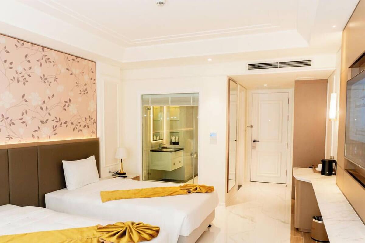 Habitación de hotel con dos camas, un espejo grande, una televisión y un baño visible a través de una puerta de cristal.
