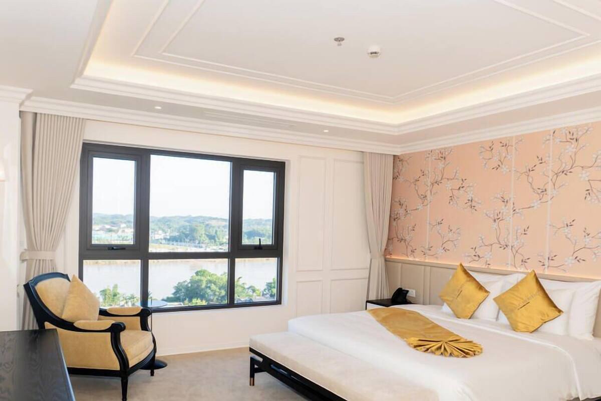 Una habitación de hotel con un ventanal grande, una cama con almohadas amarillas, una silla y un escritorio.