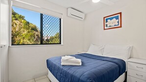 Une chambre à coucher avec un lit, des oreillers blancs, une couvre-lit bleue, une table de chevet blanche, une commode blanche, un appareil de climatisation, une fenêtre donnant sur un paysage verdoyant et un tableau encadré accroché au mur.