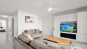 Un salon moderne avec un canapé beige, une table basse en bois, un téléviseur à écran plat fixé sur un meuble blanc et un ventilateur de plafond.