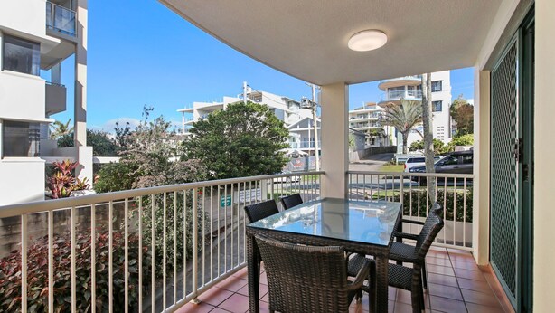 Un balcon doté d’une table en verre et de chaises en osier, donnant sur un quartier résidentiel avec de nombreux immeubles et des voitures garées.