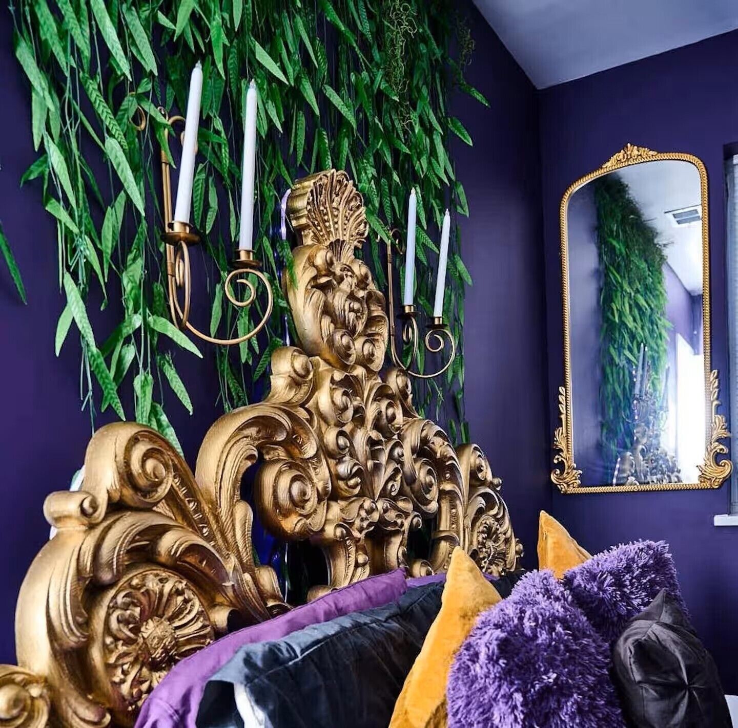 Ein Zimmer mit einem großen, verzierten goldenen Kopfteil, einem Spiegel mit goldener Rahmung und hängenden Grünpflanzen an der Wand.