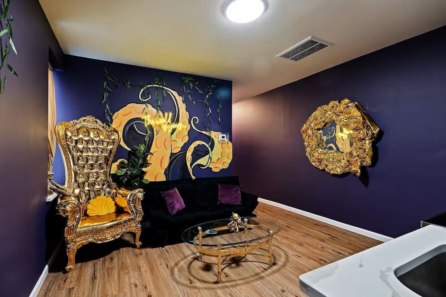 A room with a large ornate chair, a black sofa, a glass coffee table, and a gold-framed mirror on the wall.