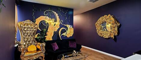 A room with a large ornate chair, a black sofa, a glass coffee table, and a gold-framed mirror on the wall.