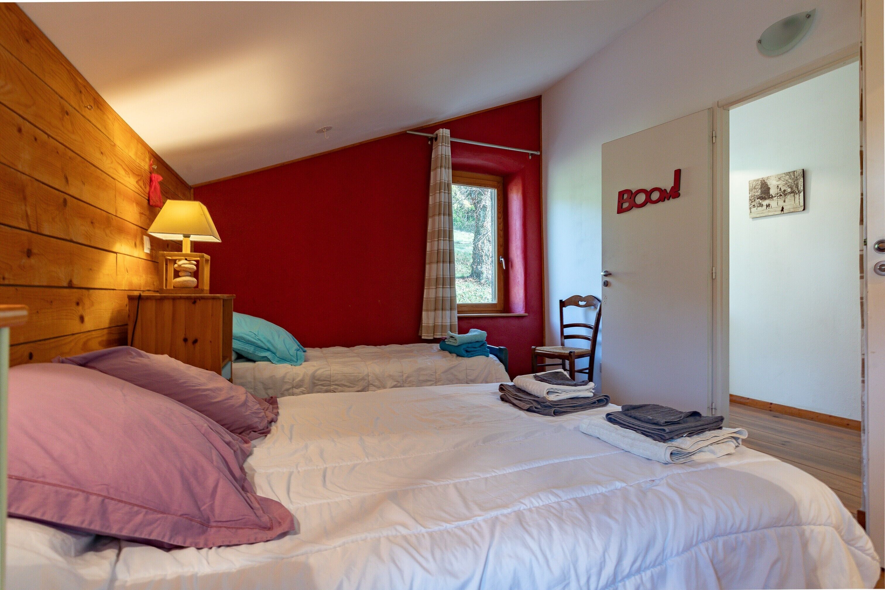 Une chambre avec deux lits, une tête de lit en bois, une fenêtre avec un rideau, une chaise et une porte avec une pancarte.