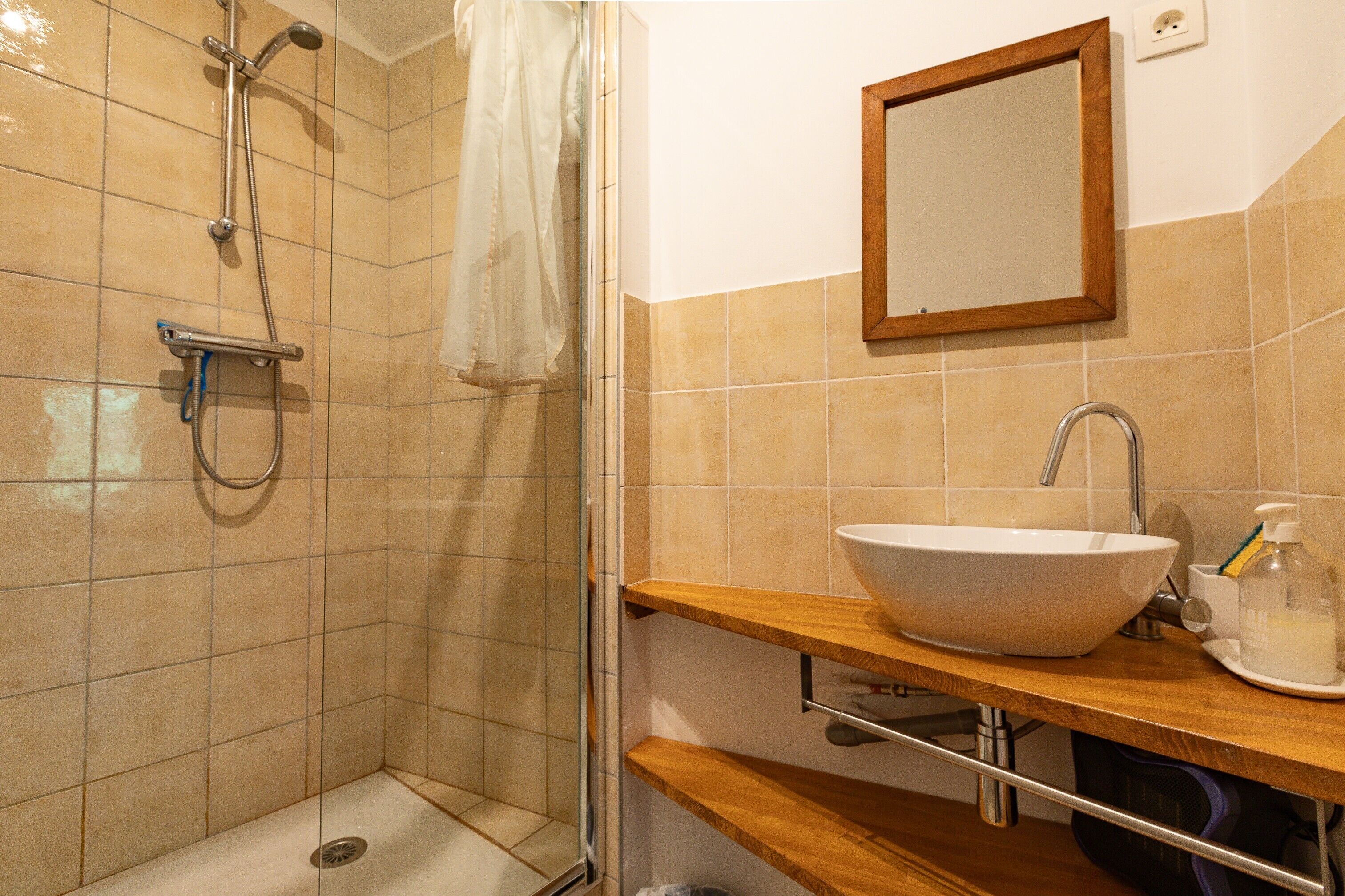 Un baño con ducha, lavamanos, espejo y estante con artículos de aseo.
