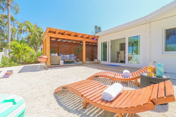 A covered outdoor seating area with wooden loungers, a grill, and a poolside lounge.