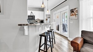 A modern kitchen with white cabinets, a black countertop, and a hanging light fixture. There is a wooden floor and a gray sofa with a woven ottoman. - Sea the Blessings (Panama City Beach)