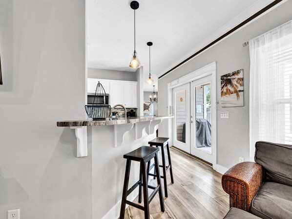 A modern kitchen with white cabinets, a black countertop, and a hanging light fixture. There is a wooden floor and a gray sofa with a woven ottoman.