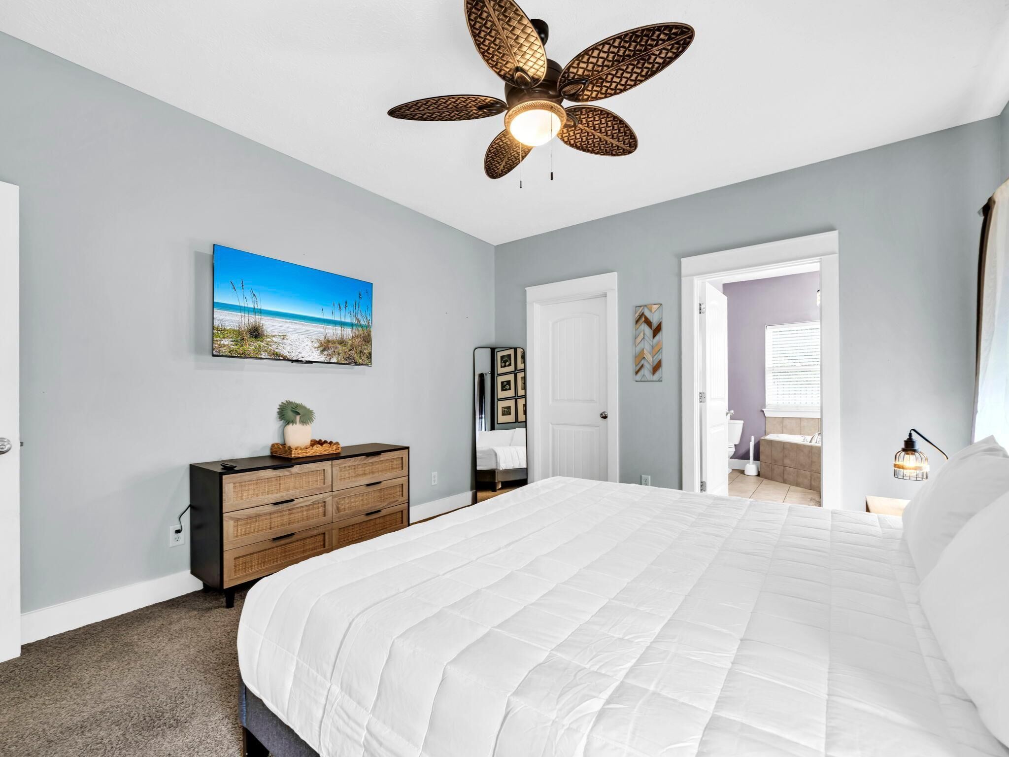 A bedroom with a large bed, a ceiling fan, a painting of a beach, a dresser, and a door leading to another room.