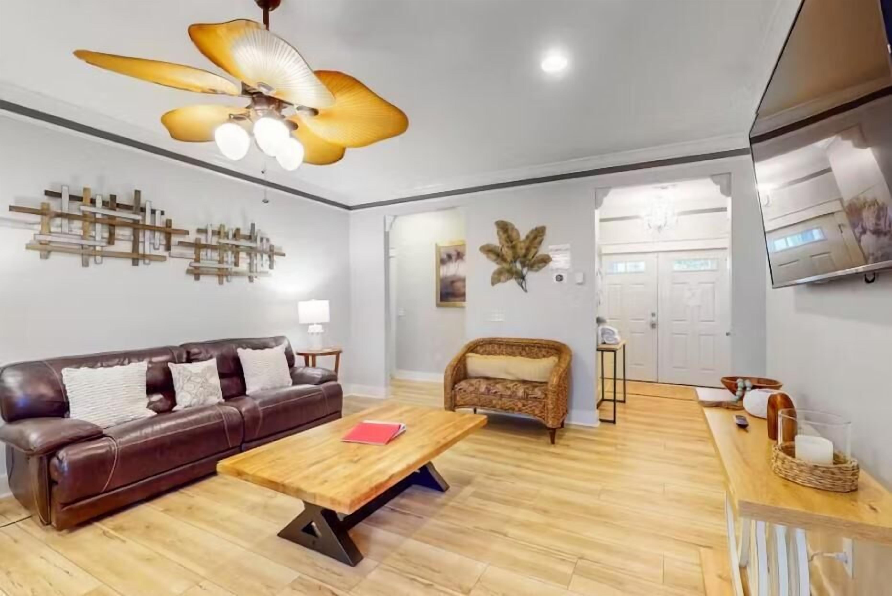 Una sala de estar moderna con un sofá de cuero marrón, una mesa de centro de madera, un sillón de mimbre y un ventilador de techo.