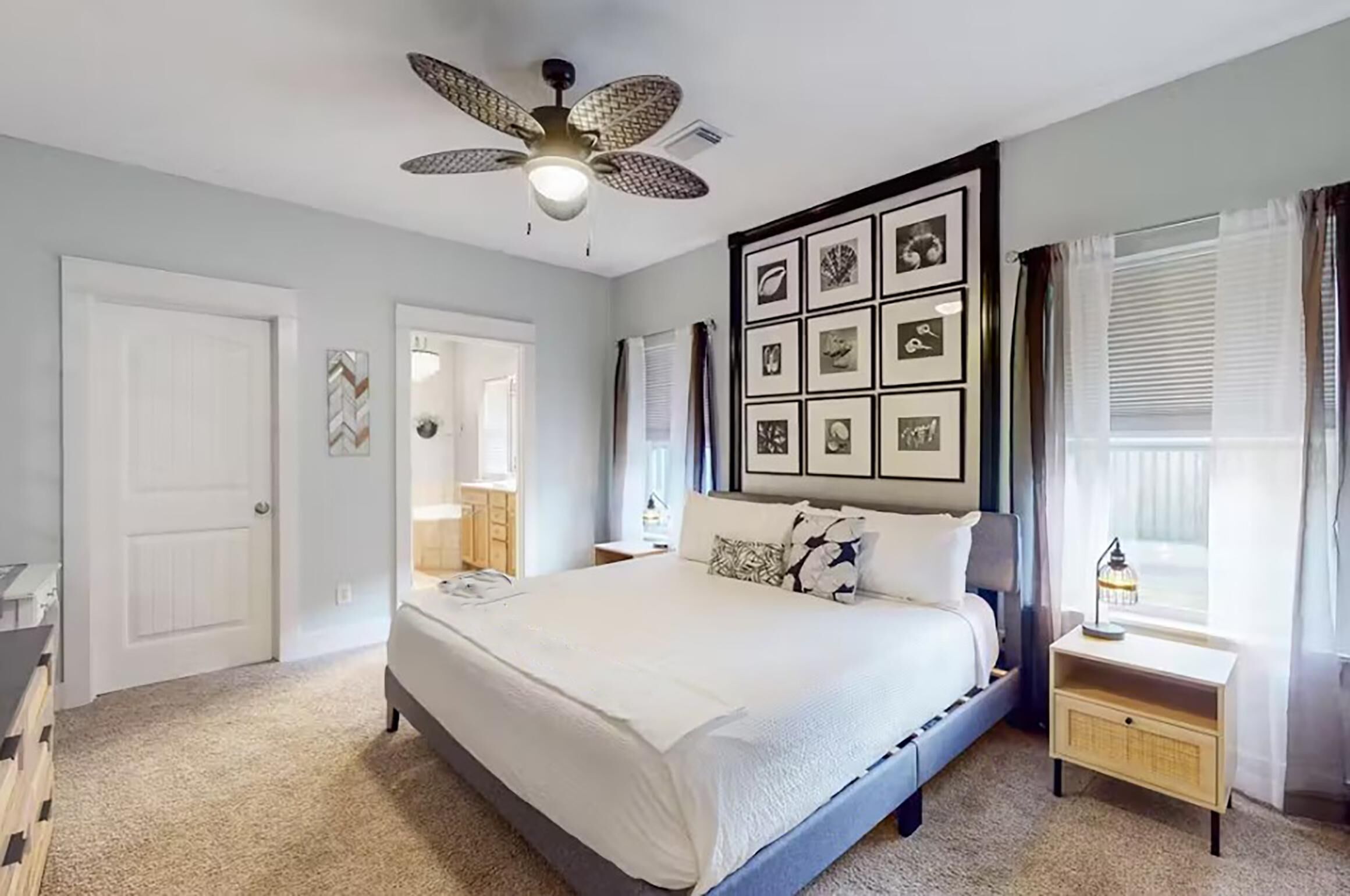 Une chambre avec un grand lit, un ventilateur de plafond, des tableaux encadrés accrochés au mur et une table de chevet avec une lampe.