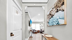 A hallway with a wooden floor, white walls, a white door, a wooden console table, and a large framed picture on the wall. - Sea the Blessings (Panama City Beach)