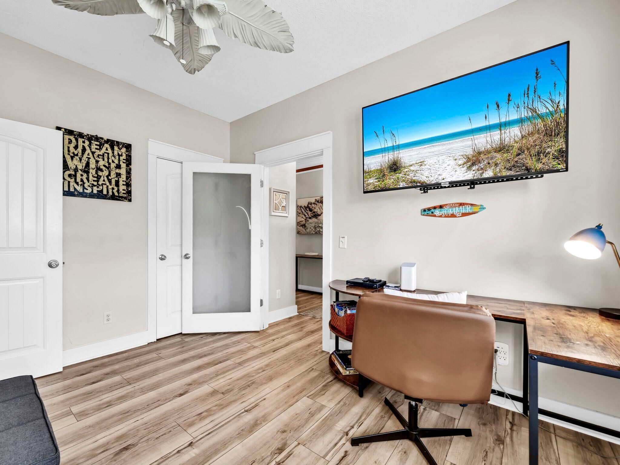 Quarto com piso de madeira, uma escrivaninha com cadeira, televisão na parede e uma porta com painel de vidro fosco.