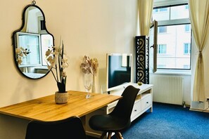 Una habitación con un escritorio de madera, televisión, espejo, un jarrón con flores y una ventana con cortinas.