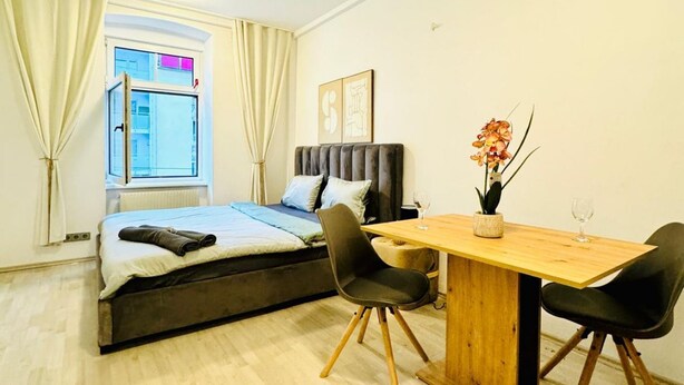 Un dormitorio con cama, ventana con cortinas, mesita de noche con un jarrón de flores y una silla.