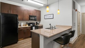 Una cocina moderna con armarios de madera oscura, un refrigerador negro, un microondas, una estufa y un lavabo. Hay dos lámparas colgantes sobre la isla, y un cartel enmarcado en la pared.