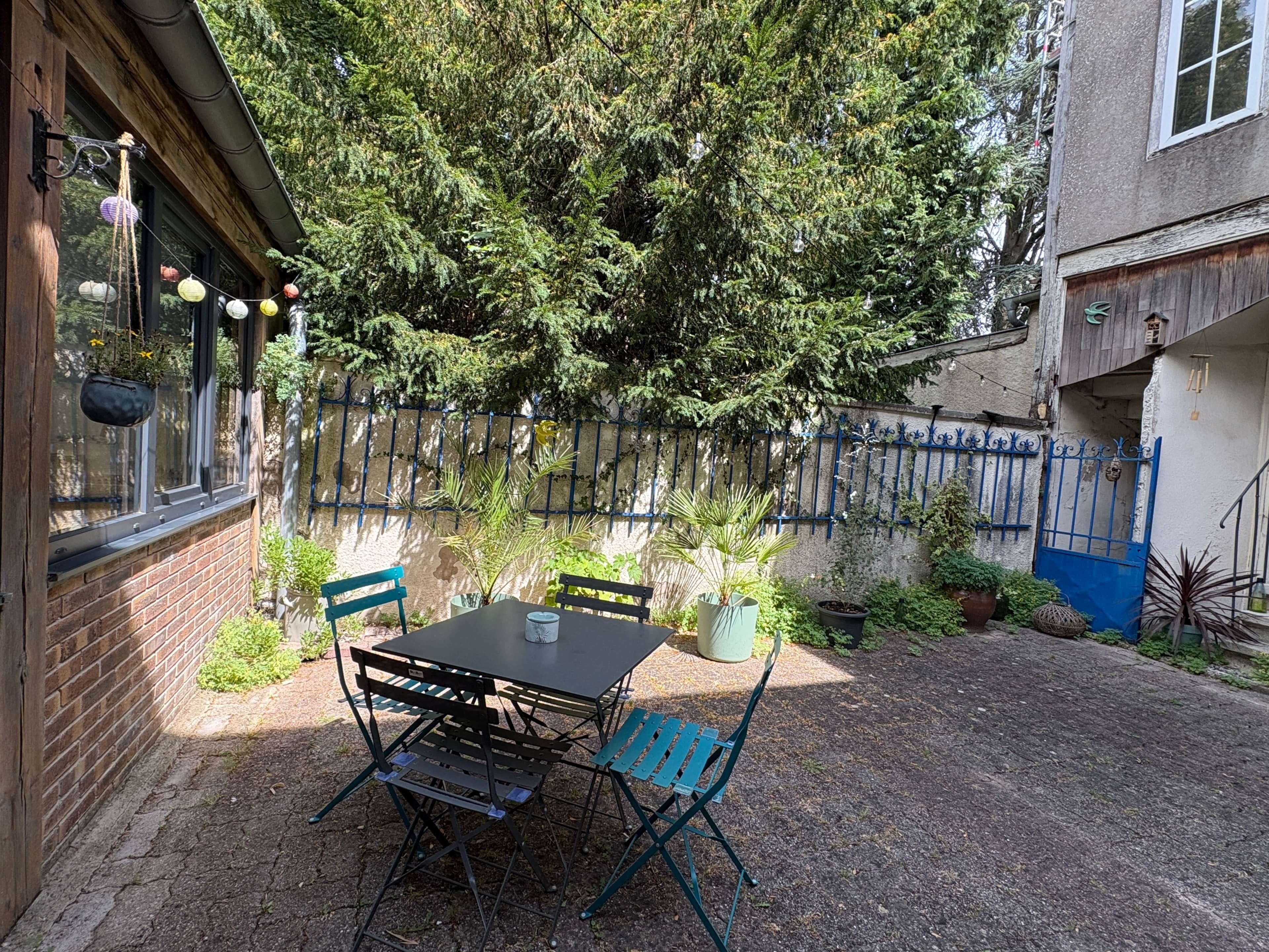Eine Terrasse im Freien mit Tisch und Stühlen, umgeben von Kübelpflanzen und einem blauen Zaun.