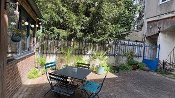 Eine Terrasse im Freien mit Tisch und Stühlen, umgeben von Kübelpflanzen und einem blauen Zaun.