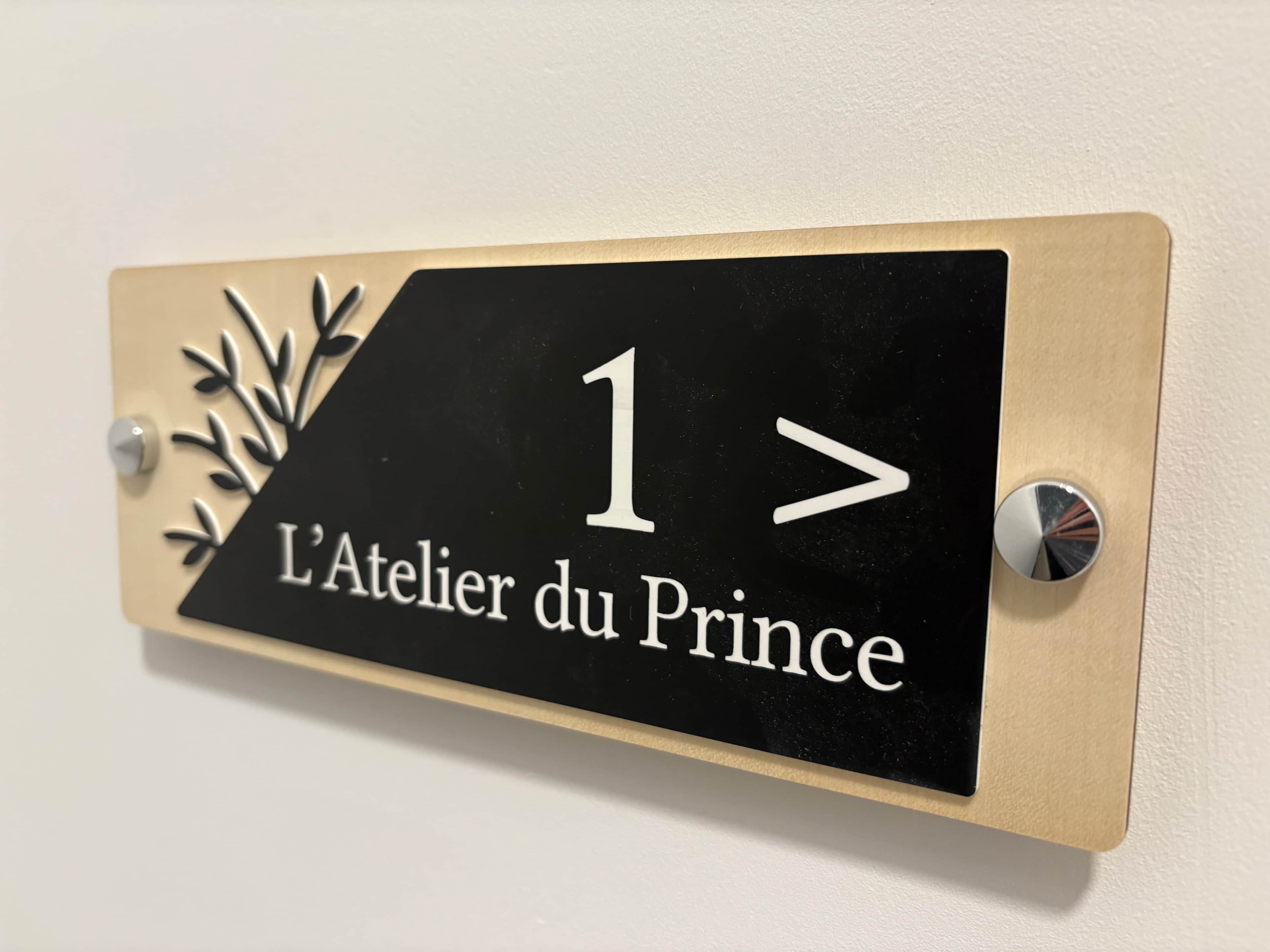 Une enseigne noire et beige, portant le numéro 1 et l’inscription « L’Atelier du Prince », est fixée à un mur.