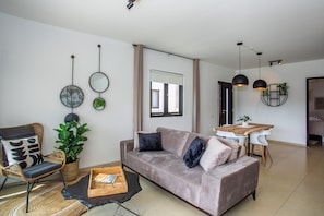 A modern living room with a sofa, a dining table, and a chair. There are plants, a mirror, and pendant lights.