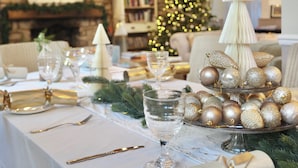 A dining table set with white tablecloth, silverware, and glassware, decorated with gold and silver ornaments, and a Christmas tree in the background.