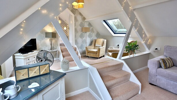 A spacious loft with a high ceiling, exposed beams, and a skylight. A staircase leads to a second level, and the room features a sofa, armchair, and a small table with decorative items.