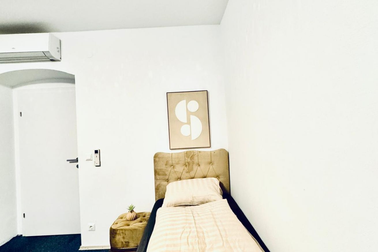 A single bed with a headboard, a wall-mounted artwork, a small round table with a plant, and a door with a handle.