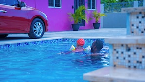 A swimming pool with two people in the water, a red car parked nearby, and a building with a purple facade in the background.