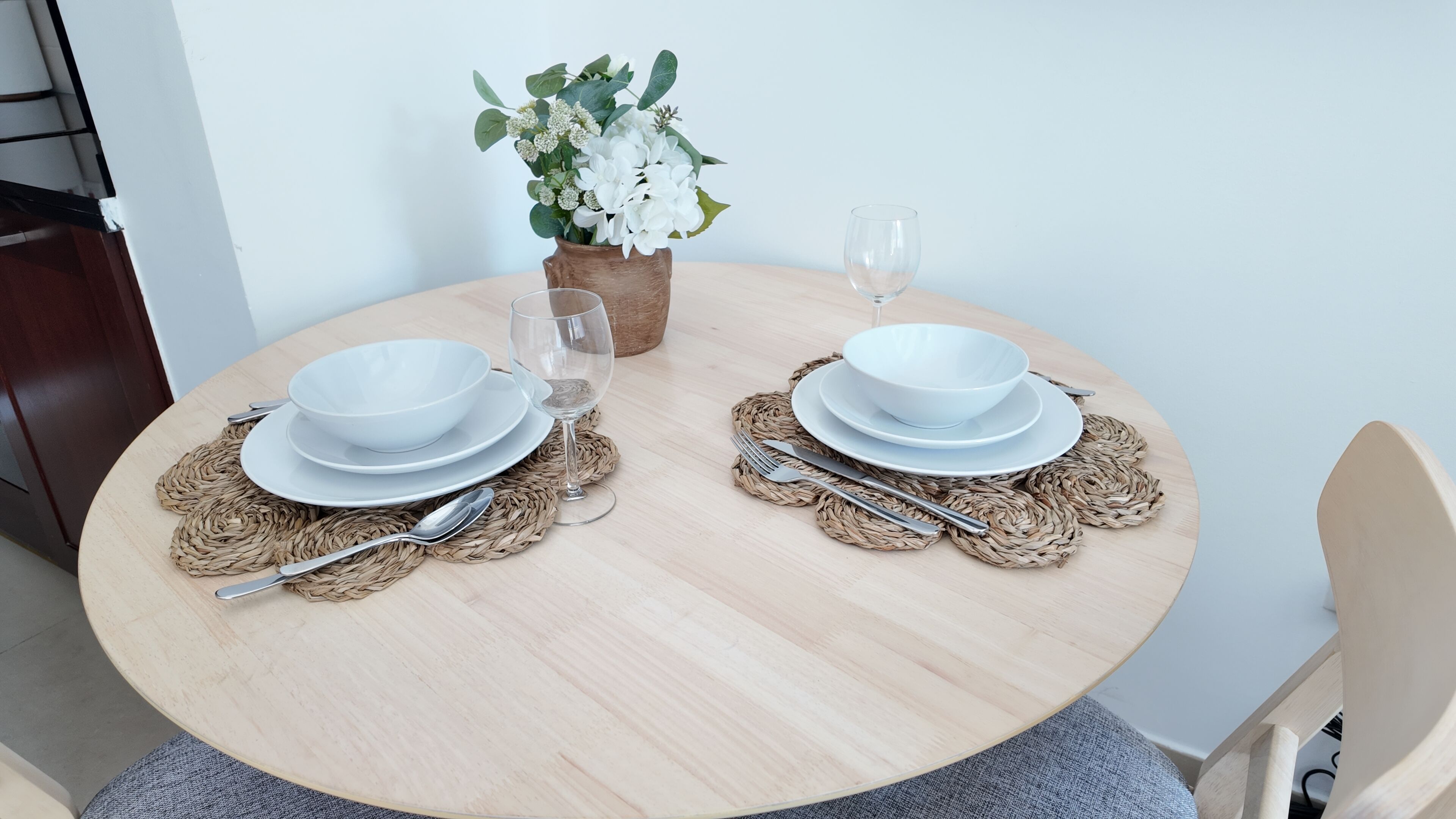 Une table de salle à manger ronde en bois, sur laquelle sont disposés deux jeux de bols, d’assiettes et de couverts blancs, posés sur des sets de table en fibres tressées. Un vase avec des fleurs blanches et un verre d’eau sont placés sur la table.