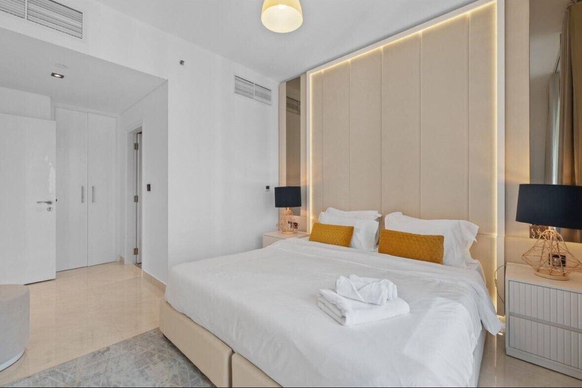 Quarto de hotel moderno com uma cama grande, mesas de cabeceira com abajures, um guarda-roupa e um banheiro visível ao fundo.