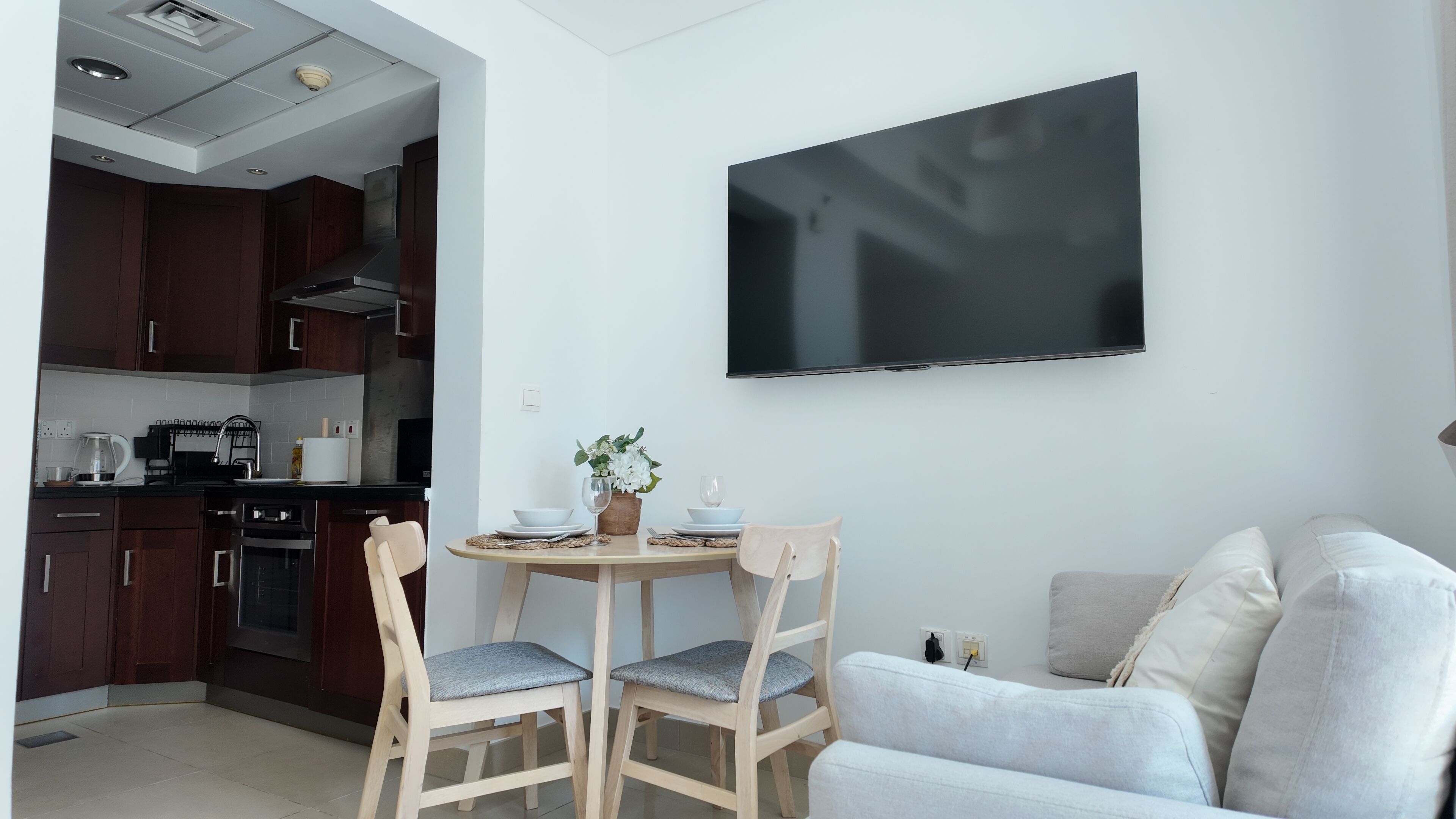 Eine moderne Küche mit dunklen Holzschränken, einem Herd und einer Kaffeemaschine. Ein Essbereich mit rundem Tisch, zwei Stühlen und einem Blumentopf. Ein Flachbildfernseher ist über dem Essbereich an der Wand befestigt.
