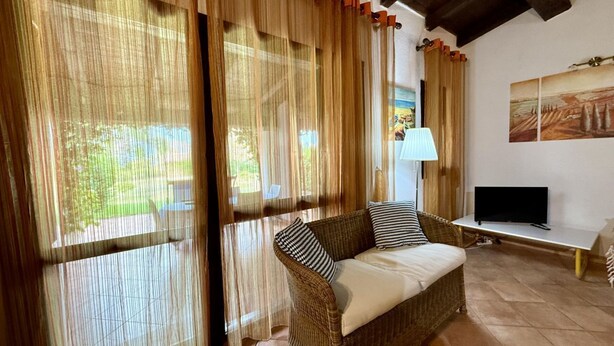 A living room with a wicker sofa, a television on a stand, and a window with sheer curtains. - Just steps from Cala D'Ambra beach and the center of San Teodoro, we offer a practical, air-conditioned three-room apartment for 4-6 people, with acourtyard and outdoor parking space. The apartment features a living room with a sofa bed and TV, a fully e (San Teodoro)