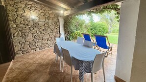 A covered patio with a stone wall, a table with chairs, and a view of greenery. - Just steps from Cala D'Ambra beach and the center of San Teodoro, we offer a practical, air-conditioned three-room apartment for 4-6 people, with acourtyard and outdoor parking space. The apartment features a living room with a sofa bed and TV, a fully e (San Teodoro)