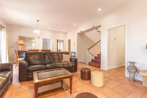 A spacious living area with a leather sofa, a glass coffee table, a dining table with chairs, a staircase, and a vase on the floor.