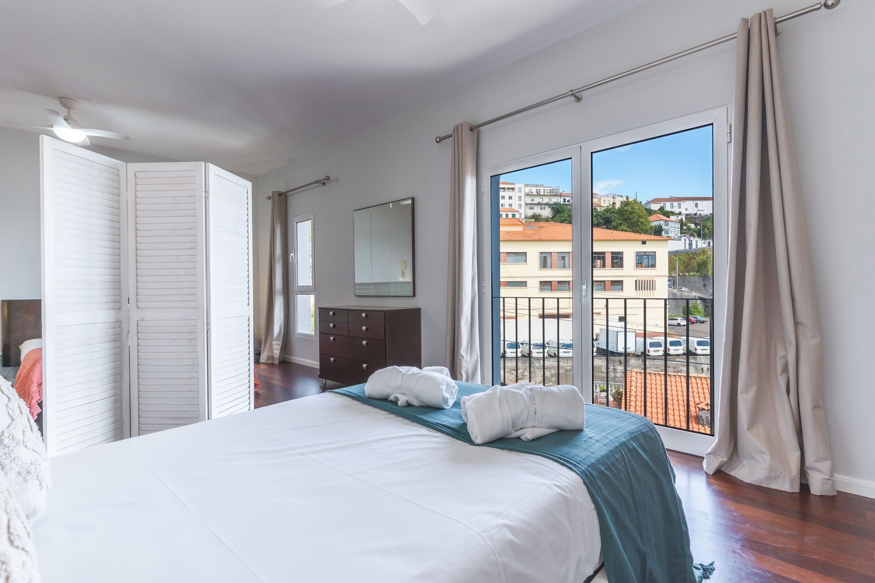 A bedroom with a large bed, a dresser, a mirror, and a balcony with a view of buildings.
