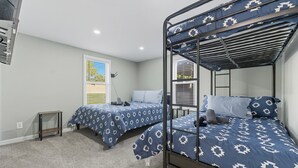 Une chambre avec deux lits superposés, chacun recouvert d’une courtepointe bleue et blanche à motifs, une petite table de chevet en bois et une fenêtre avec des stores.