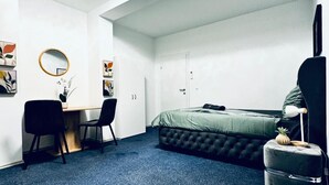 Una habitación con una cama, un escritorio, dos sillas, un espejo redondo y una planta pequeña sobre un taburete.