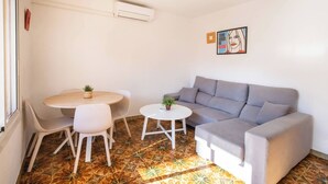A small, modern living space with a grey sofa, a round coffee table, and two white chairs. There is a small plant on the coffee table and another on the sofa. The room has a patterned floor and a wall-mounted air conditioning unit. - Apartment in Gracia A34PAA1127 (Barcelona)