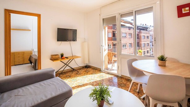A modern living room with a sofa, a small table with a plant, a TV mounted on the wall, and a balcony with a view of buildings. - Apartment in Gracia A34PAA1127 (Barcelona)