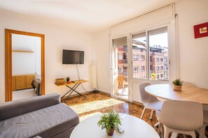 Un salon moderne avec un canapé, une petite table sur laquelle est posée une plante, un téléviseur fixé au mur et un balcon offrant une vue sur les bâtiments.