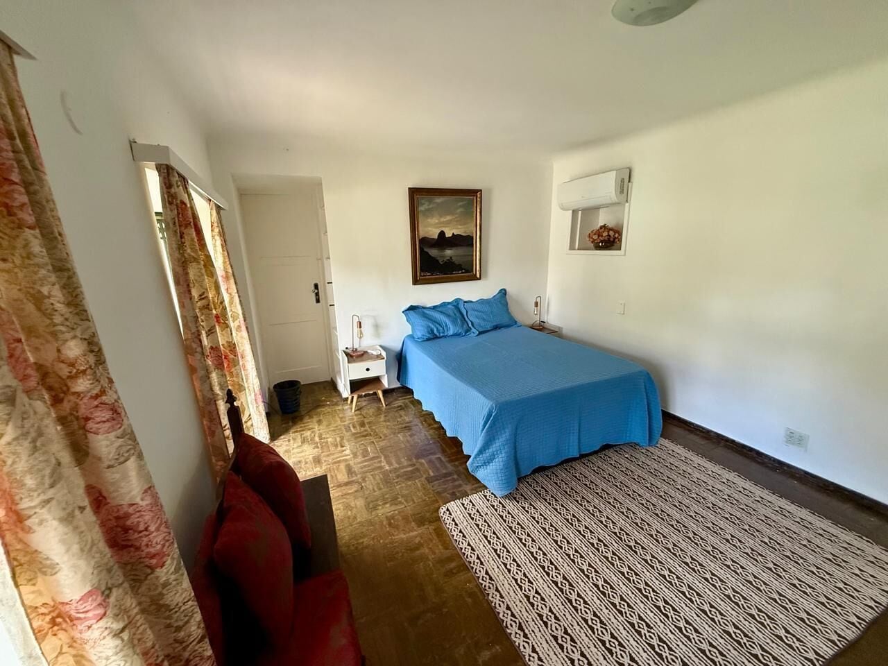 Een slaapkamer met een bed, nachtkastje, rode stoel, een kamerbreed tapijt en een schilderij aan de muur.