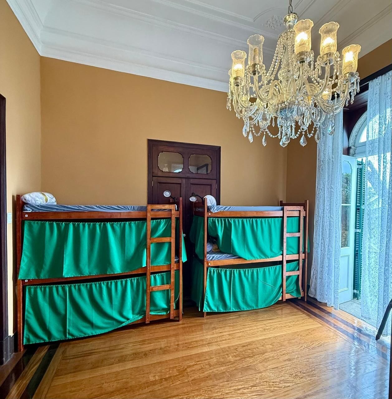 A room with two bunk beds, a wooden door with glass panels, a chandelier, and a view through a window with curtains.