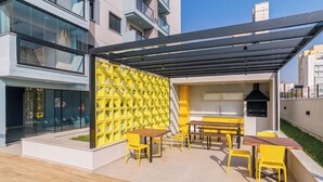 An outdoor patio with yellow chairs and tables, a grill, and a yellow geometric wall. - Apto no Butant c Academia e pr Ximo ao Metr (São Paulo)