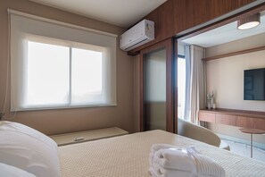 A hotel room with a bed, a bedside table, a desk, a chair, a large window, an air conditioning unit, and a mirror. - Apto no Butant c Academia e pr Ximo ao Metr (São Paulo)
