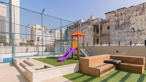 A rooftop playground with a slide and a play structure, surrounded by a fence and a building with graffiti. - Apto no Butant c Academia e pr Ximo ao Metr (São Paulo)