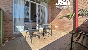 Een terras met twee stoelen en een tafel, een schuifdeur die naar een kamer leidt, en een bakstenen muur met een bord waarop Jindabyne Ski Accommodation staat.