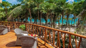 Eine Holzterrasse mit Korbmöbeln und Blick auf einen Strand mit Palmen und türkisblauem Wasser.