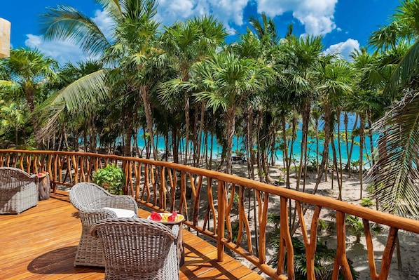 A wooden deck with wicker furniture and a view of a beach with palm trees and turquoise water.